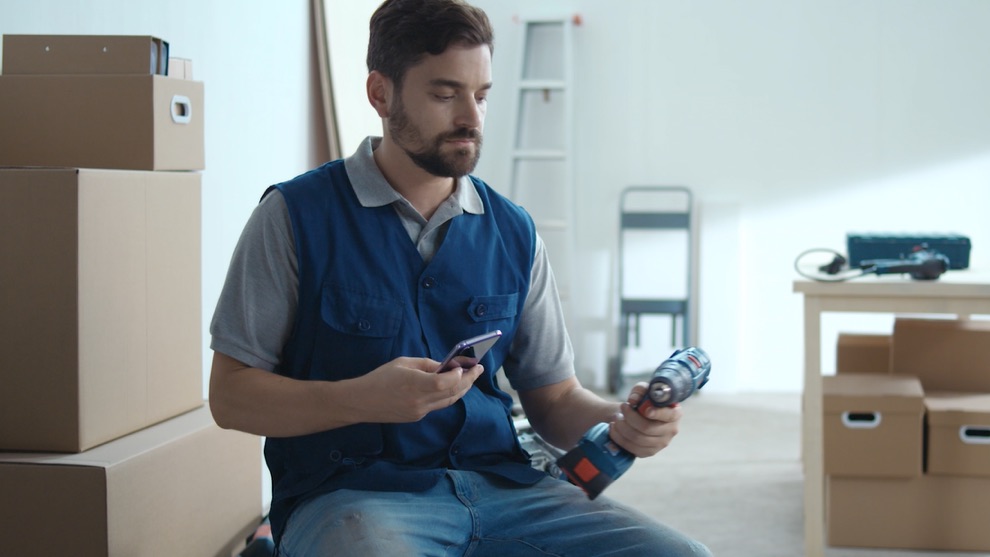 Giới thiệu BeConnected  Một người đàn ông mặc áo vest màu xanh đang cầm một chiếc tua vít không dây Bosch và một chiếc điện thoại di động.