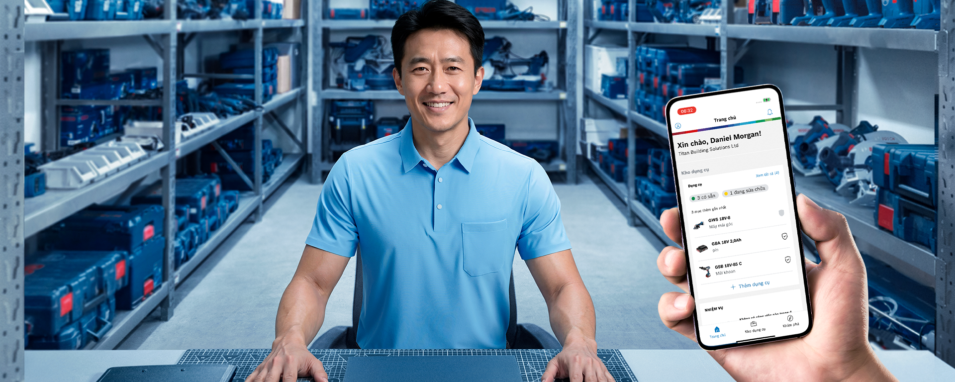 Một người đàn ông mặc áo sơ mi xanh ngồi tại một chiếc bàn trong một nhà kho đầy dụng cụ và thùng hàng.