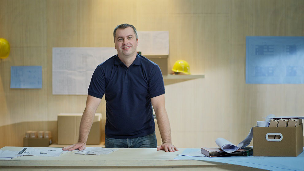 Một người đàn ông mặc quần áo màu xanh dương đang mỉm cười đứng bên bàn, xung quanh là bản vẽ và các tập tài liệu. Phía sau là những chiếc mũ bảo hiểm màu vàng và các bản kế hoạch.