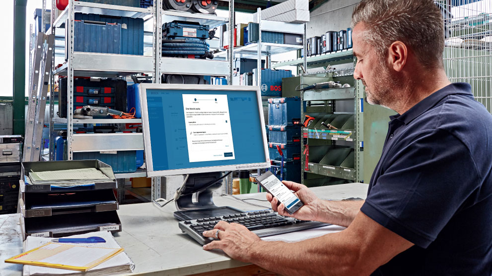 Một người đàn ông đang làm việc trên máy tính trong khi cầm điện thoại thông minh. Phía sau là những kệ đựng hộp dụng cụ và vật liệu.