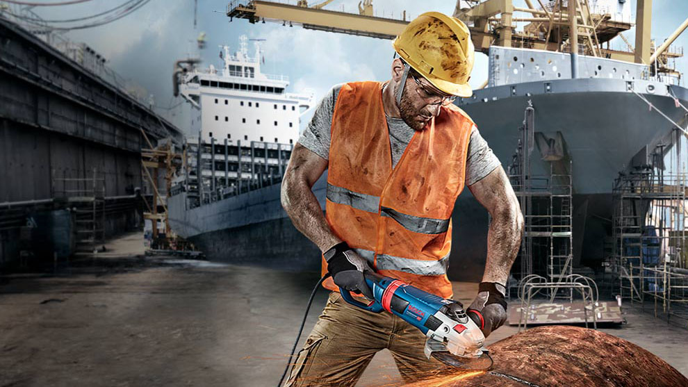 Một công nhân đội mũ bảo hiểm đang cắt kim loại bằng máy Bosch màu xanh.