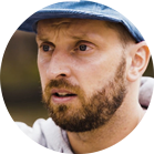 Một người đàn ông có râu đội mũ xanh và nhìn nghiêm túc.