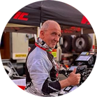 Một người đàn ông đội mũ bảo hiểm và mặc bộ đồ đua giơ ngón tay cái lên trước một cái lều và các công cụ.