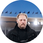 Một người đàn ông có râu và áo khoác đen đứng dưới ánh đèn sân.