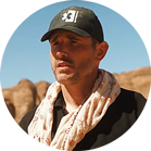 Một người đàn ông đội mũ đen và quàng khăn đứng trước một cảnh quan đá.