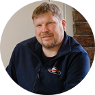 Một người đàn ông mỉm cười với tóc ngắn đang mặc một chiếc áo len màu xanh.